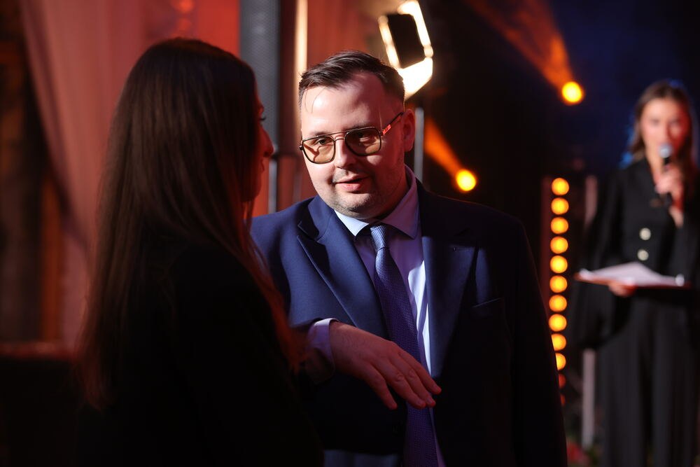 Rozmowa na evencie — gość na pierwszym planie, prowadząca z mikrofonem w tle